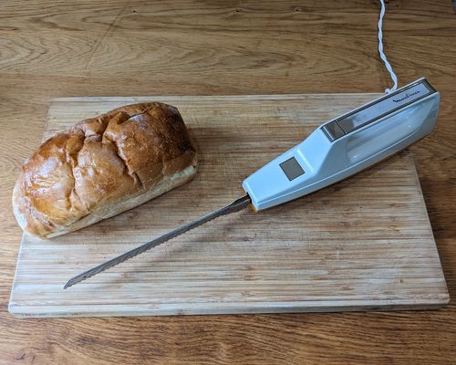 Le couteau électrique placé sur une planche à découper en bois, à côté d’un pain de mie pas encore découpé