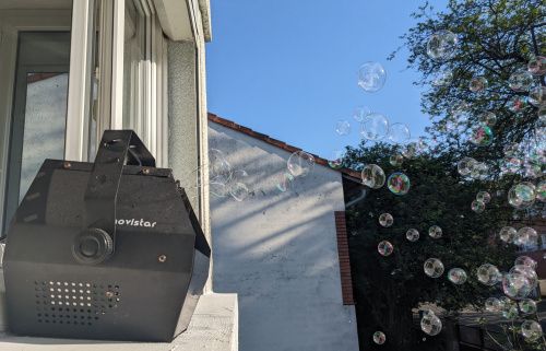 Machine à bulles en fonctionnement sur un appui de fenêtre, bulles devant la maison voisine et les arbres
