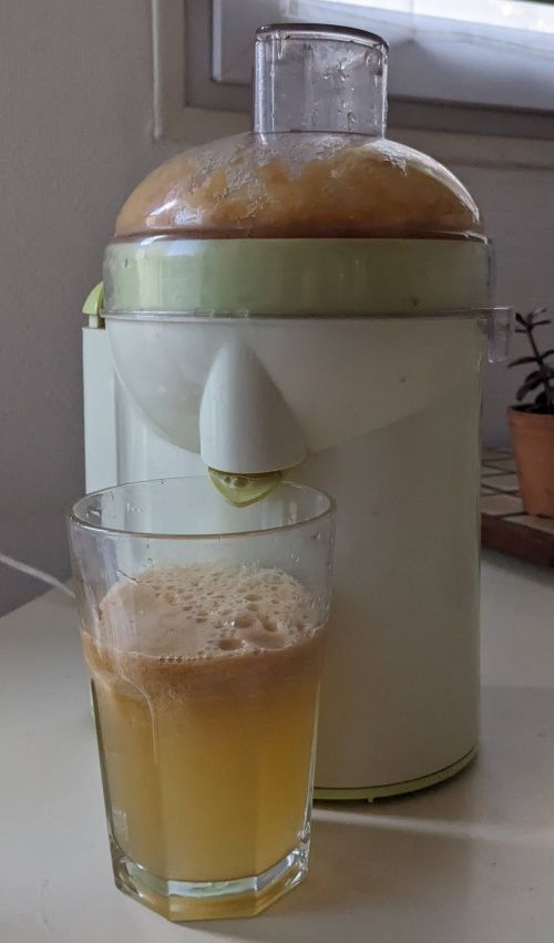 Le verre rempli de jus doré surmonté de mousse, devant la centrifugeuse dont le couvercle est plein de pulpe